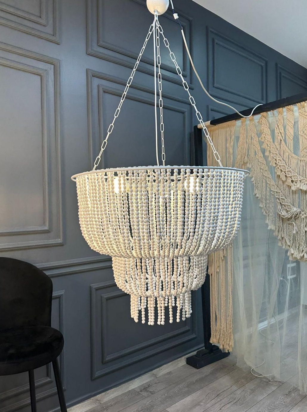 “Handmade white beaded chandelier with three lights, shown hanging in a modern boho living room. Large pendant light made of painted wooden beads.”
